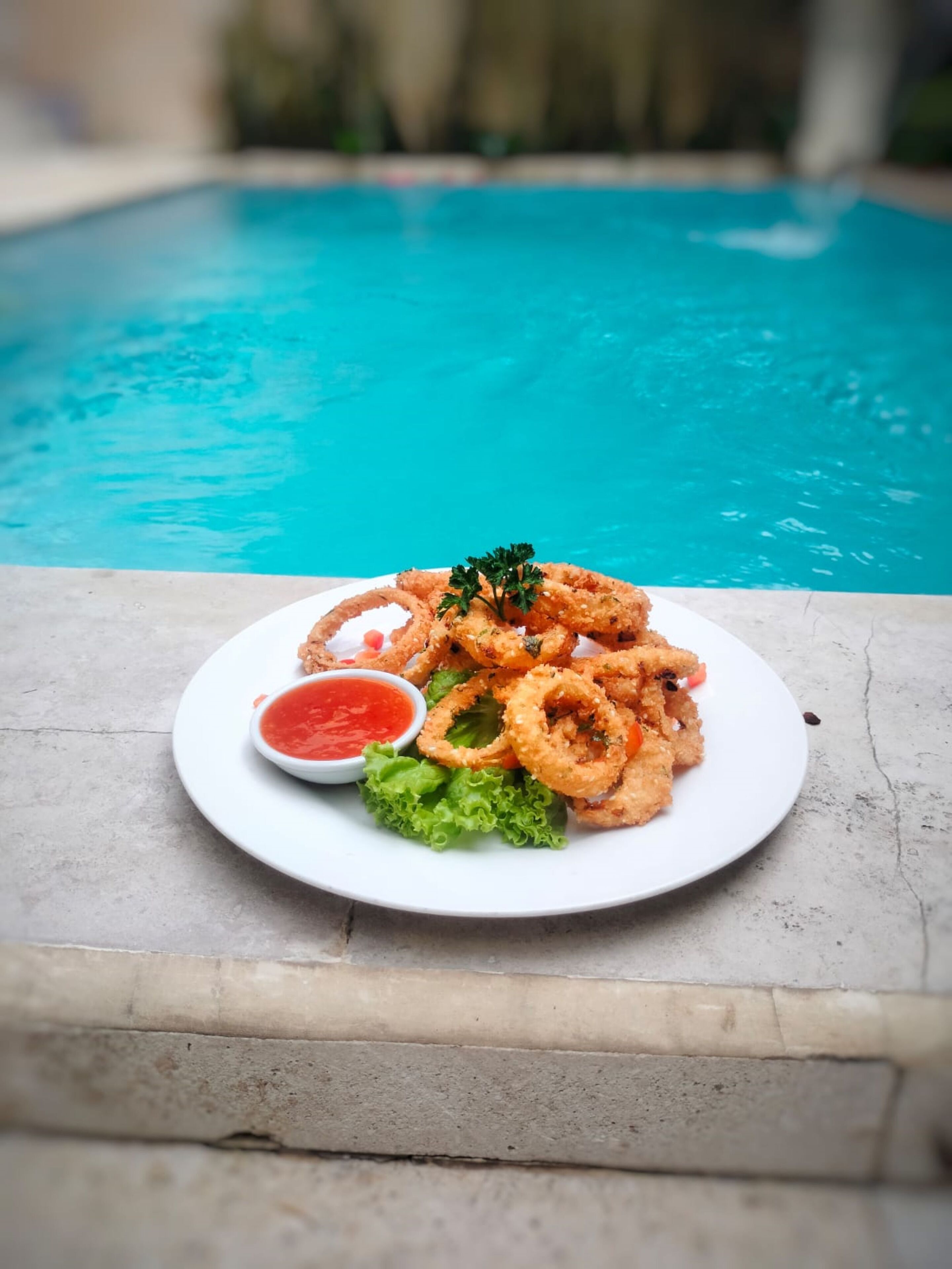 Gebratene Calamare auf einem Teller, dazu rote Soße und Salat, mit Blick auf einen Pool.