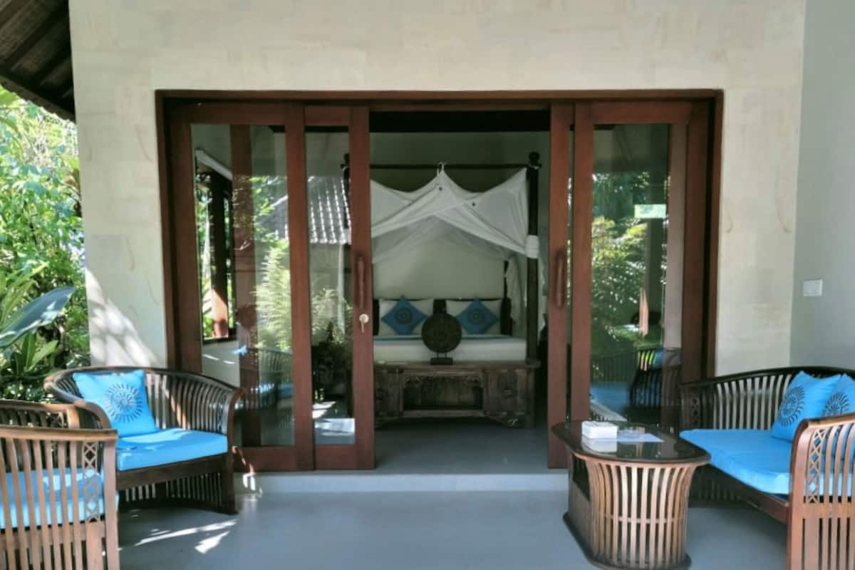 A room with a glass door leading to a bed with a canopy, two wicker chairs with blue cushions, and a small table.