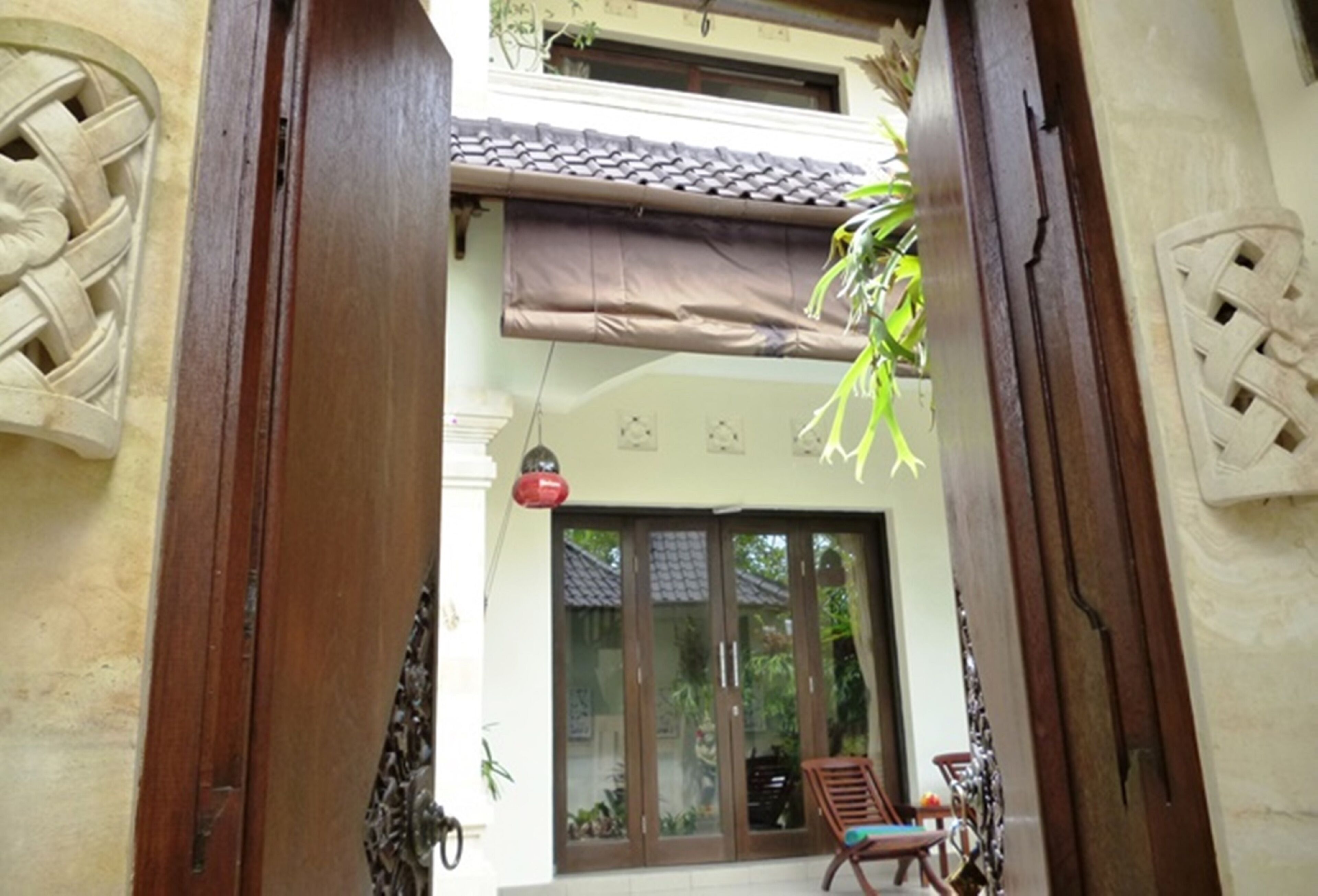 A traditional wooden gate with intricate carvings, a tiled roof, and a sliding glass door leading to a balcony with a chair and a hanging plant.