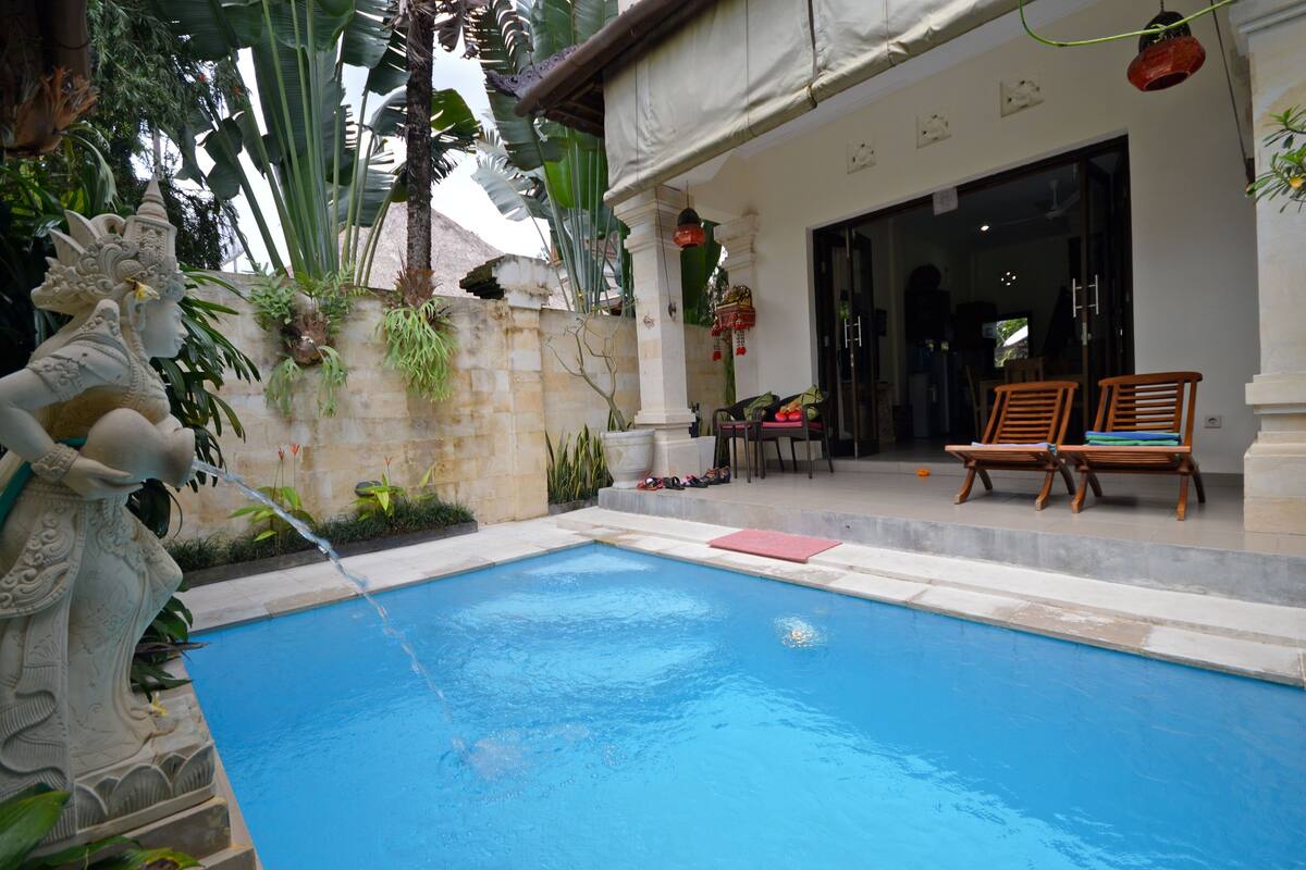 A swimming pool with a statue, a covered patio with chairs, and a stone wall with plants.