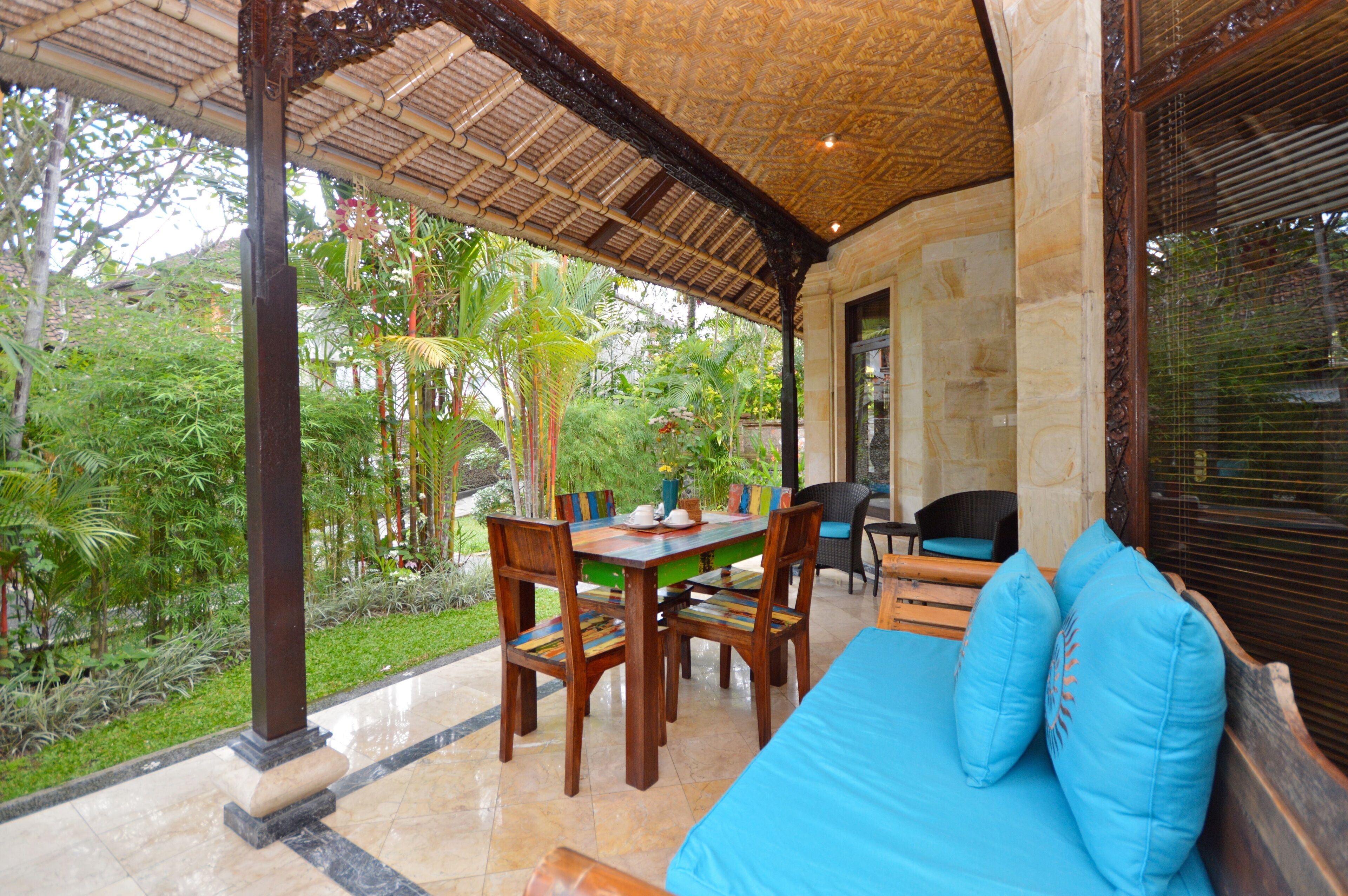 A covered outdoor dining area with a table and chairs, a cushioned seating area, and a view of lush greenery.