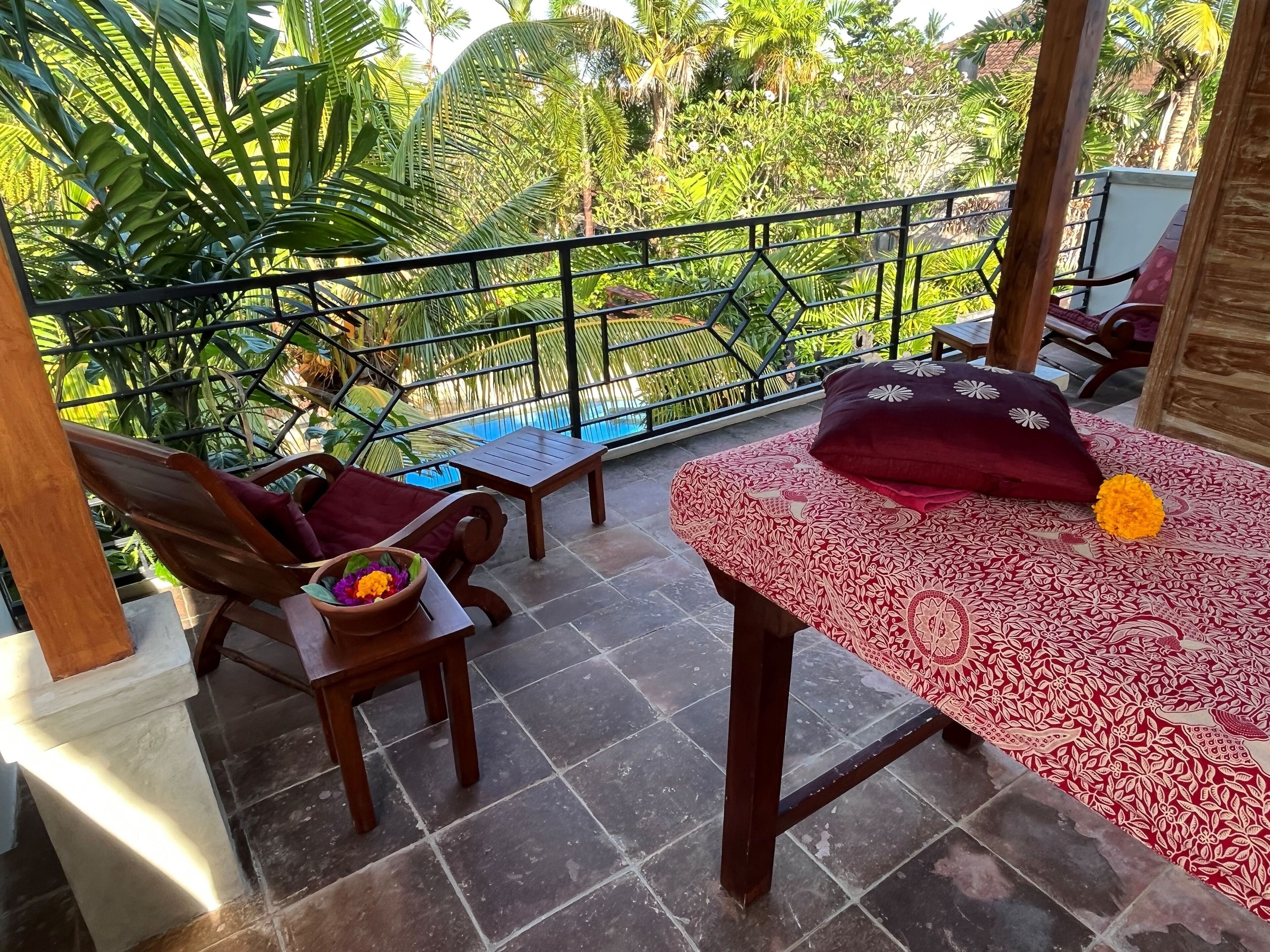 A balcony with a wooden table, a chair, and a cushioned bench. There is a view of lush greenery and a pool in the background.