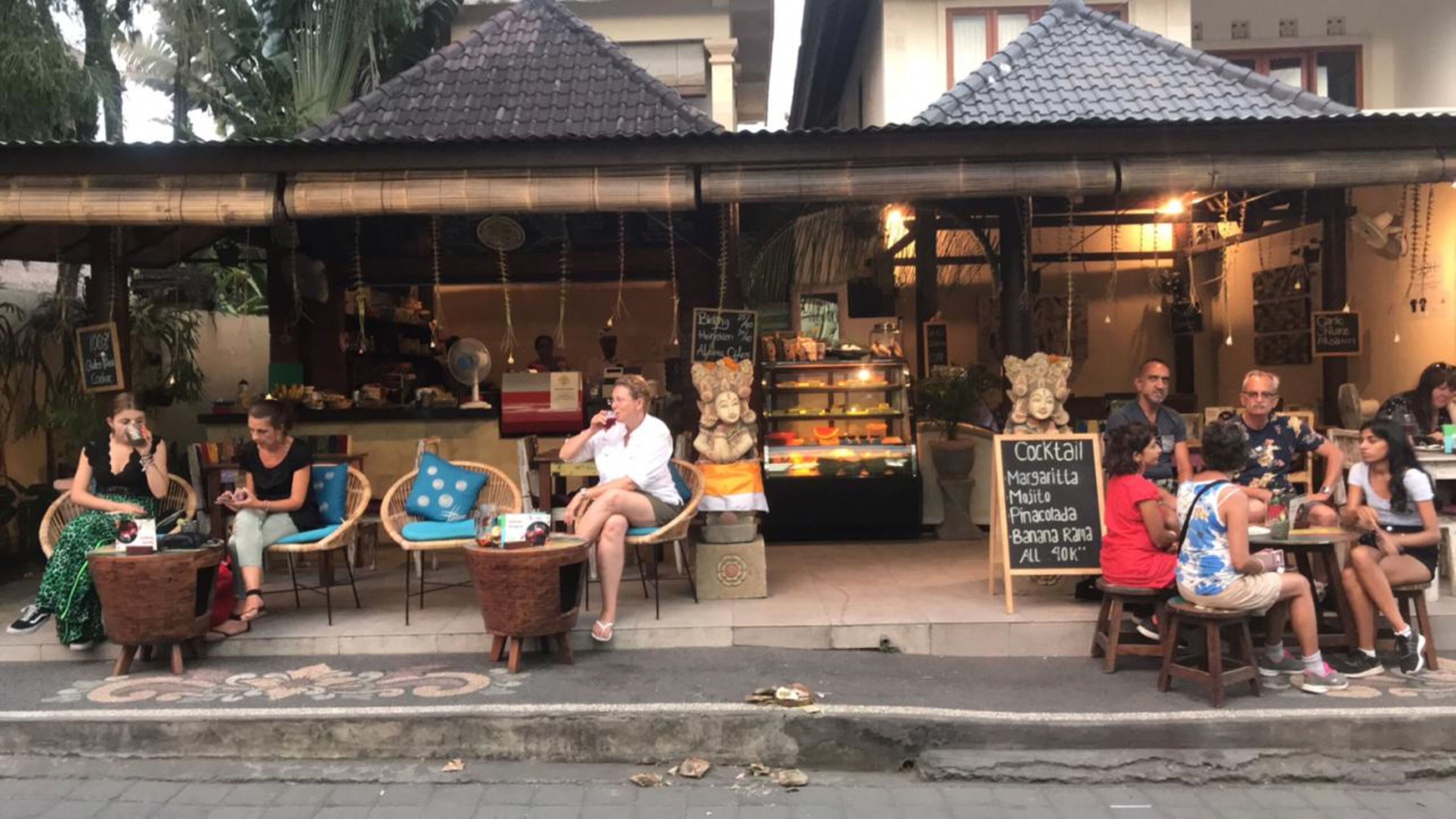 An outdoor café with wicker chairs and tables, a signboard with drink options, and a building with a tiled roof in the background.