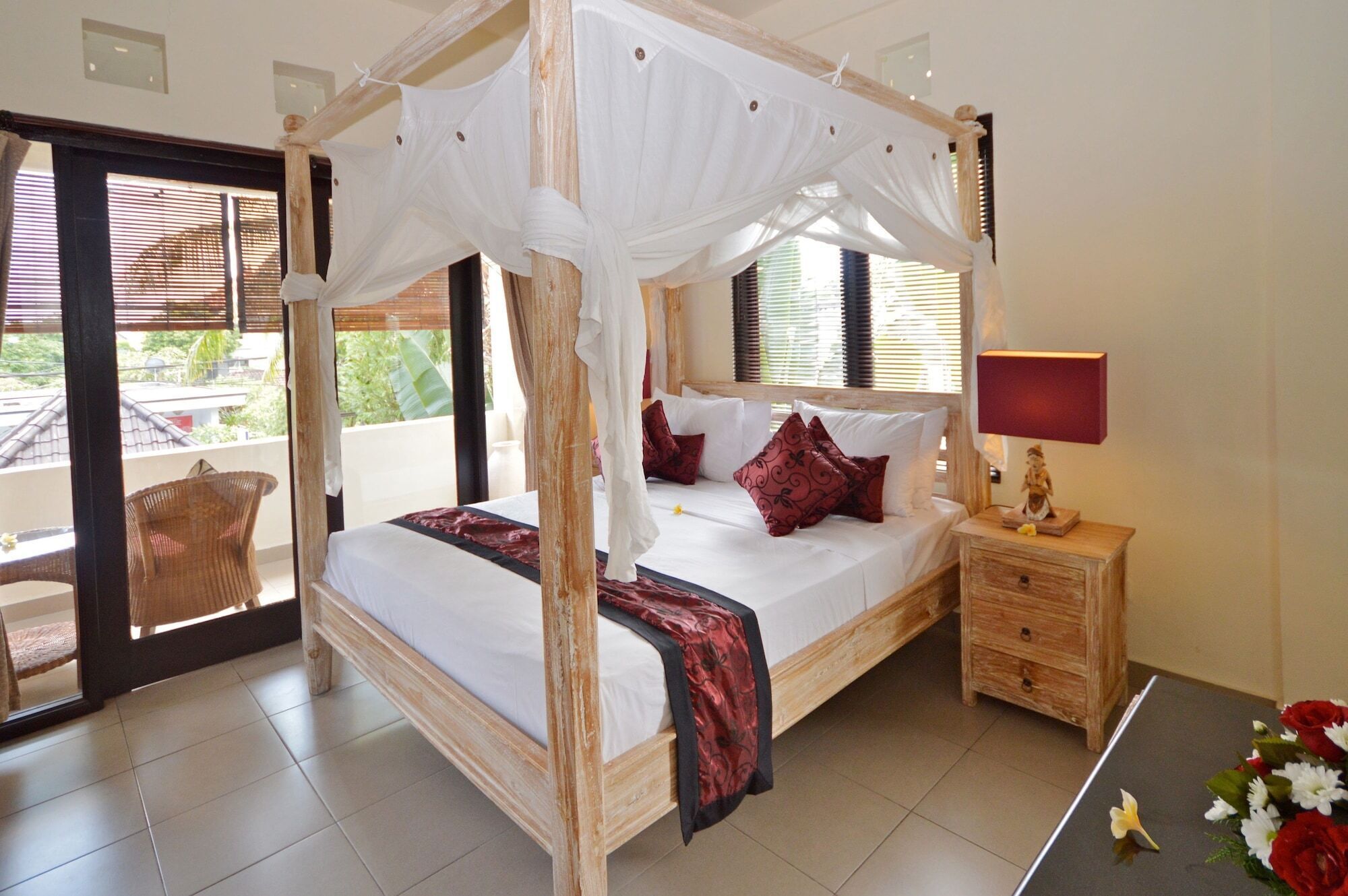 A four-poster canopy bed with a wooden frame, white bedding, and red pillows. There is a nightstand with a lamp and a small statue. The room has a balcony with a wicker chair and a view of greenery.