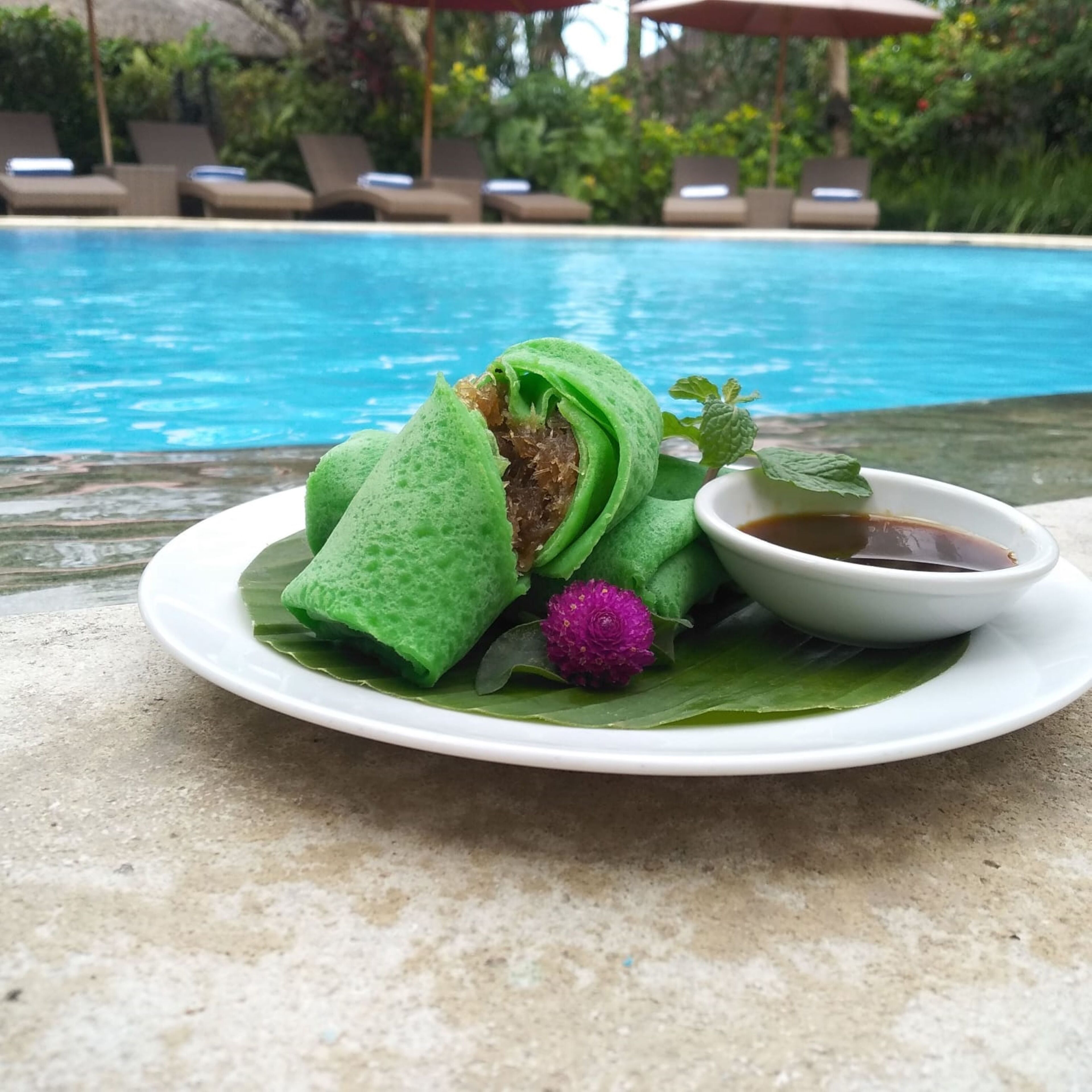 A plate with a green dessert, a purple flower, and a small bowl of sauce by a poolside.