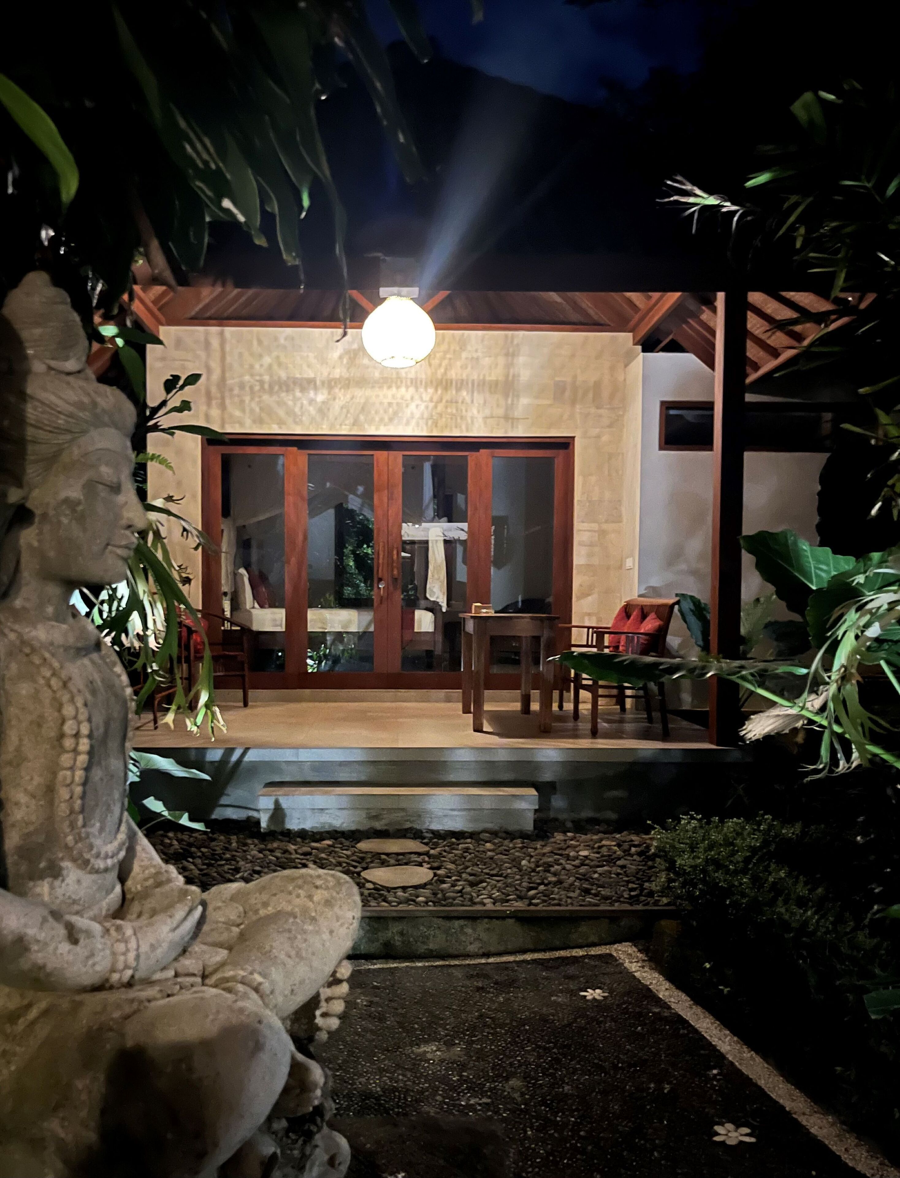 A covered outdoor seating area with a table and chairs, surrounded by plants and a stone statue.