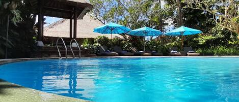 A clear blue pool with lounge chairs and blue umbrellas, surrounded by lush greenery and a thatched-roof structure.