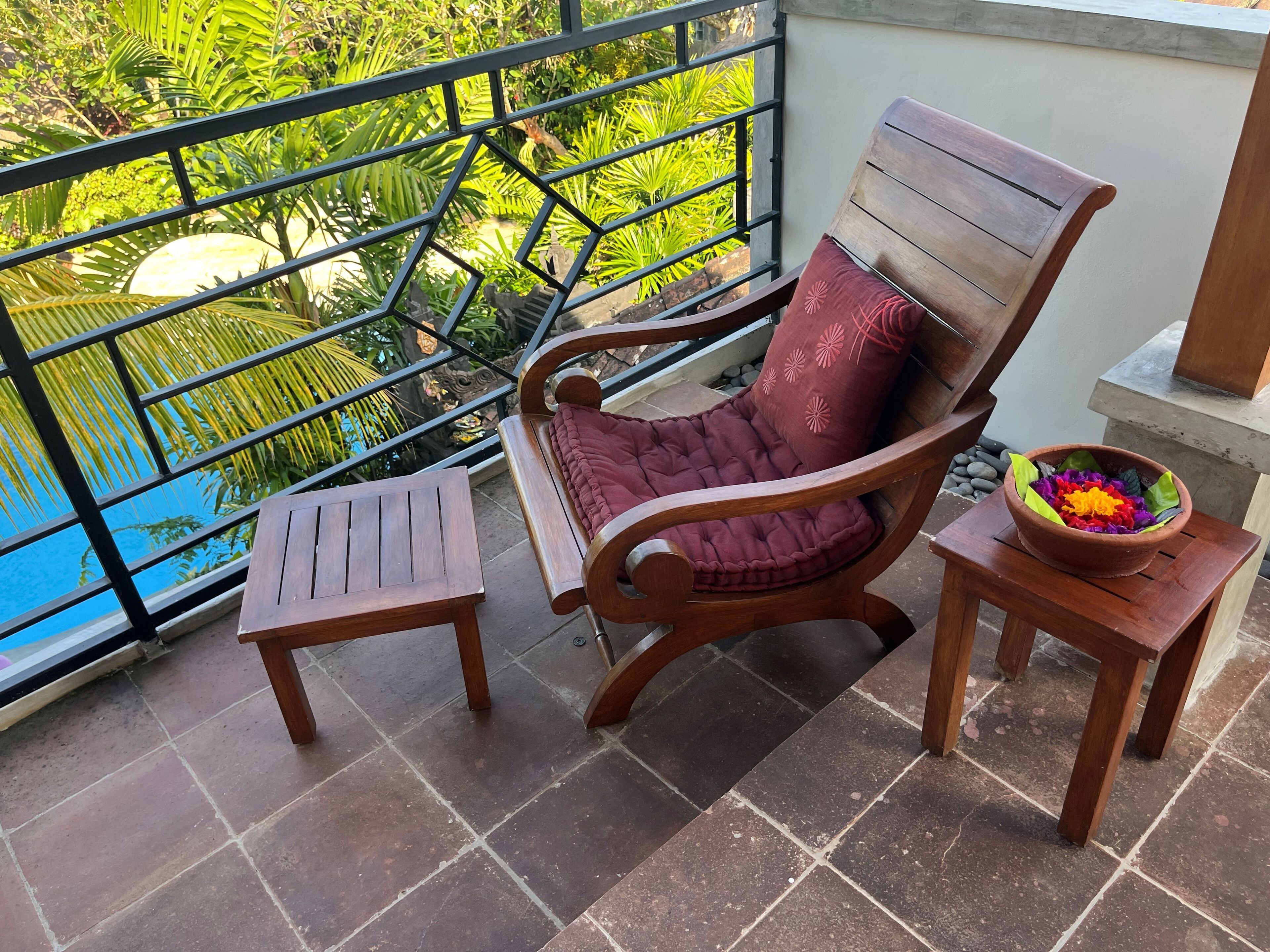 A wooden chair with a cushioned seat and a red pillow, placed on a tiled balcony with a view of a swimming pool and lush greenery.