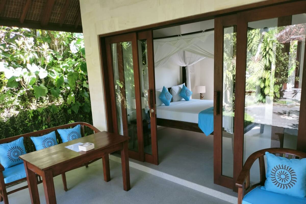 A room with a bed, a table, and chairs. There is a view of a garden through the glass doors.