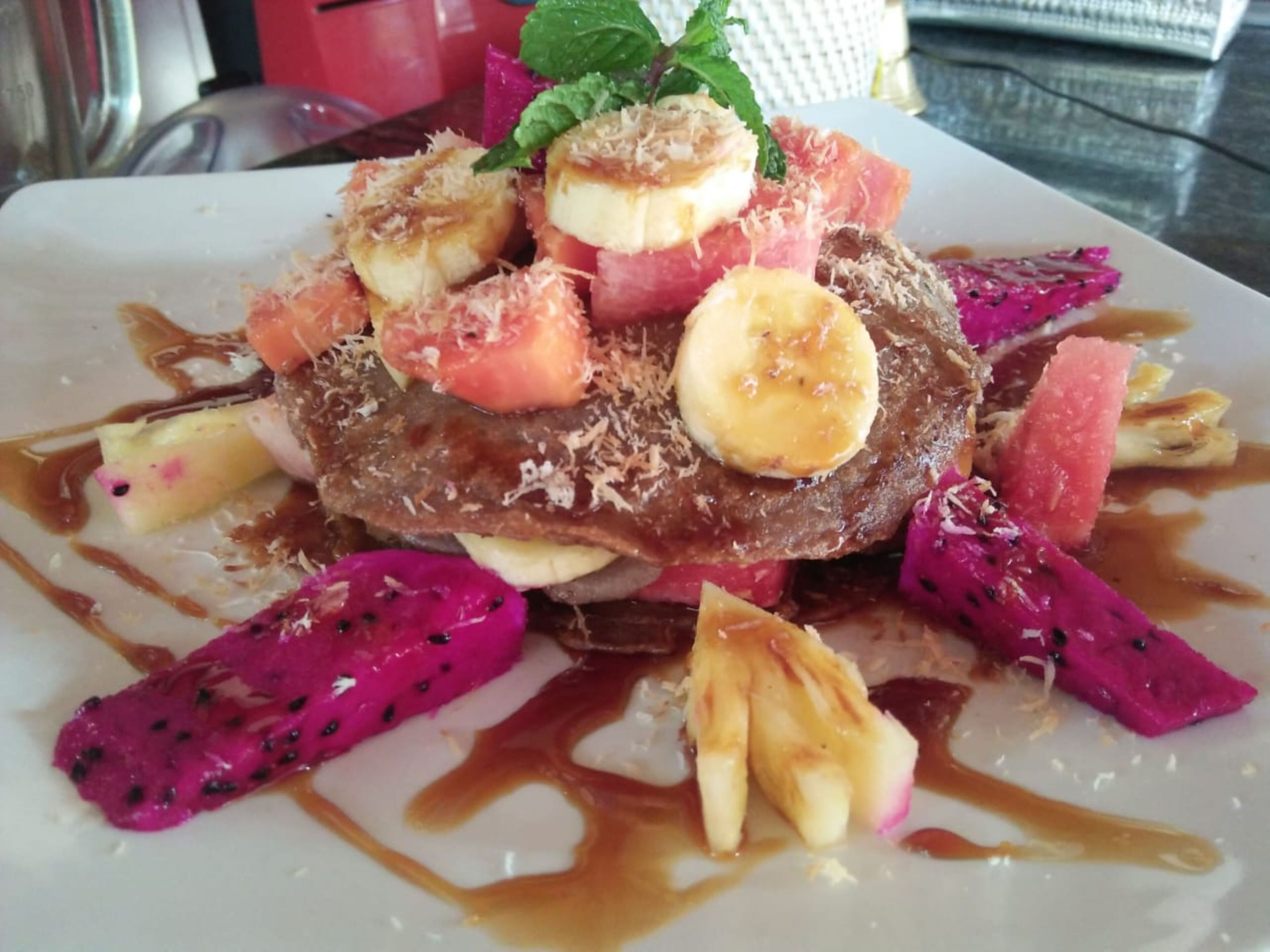 Un plato de postre con tortitas, plátanos, pitahaya y salsa de caramelo.