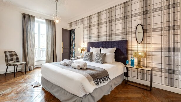 A modern hotel room with a large bed, plaid wallpaper, a wooden floor, a chair, and a nightstand with lamps. - Sweett | Rivoli II - Three Bedroom Apartment, Sleeps 6 (Paris)