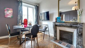 A dining area with a fireplace, a TV mounted on the wall, and a window with curtains. - Sweett | Rivoli II - Three Bedroom Apartment, Sleeps 6 (Paris)