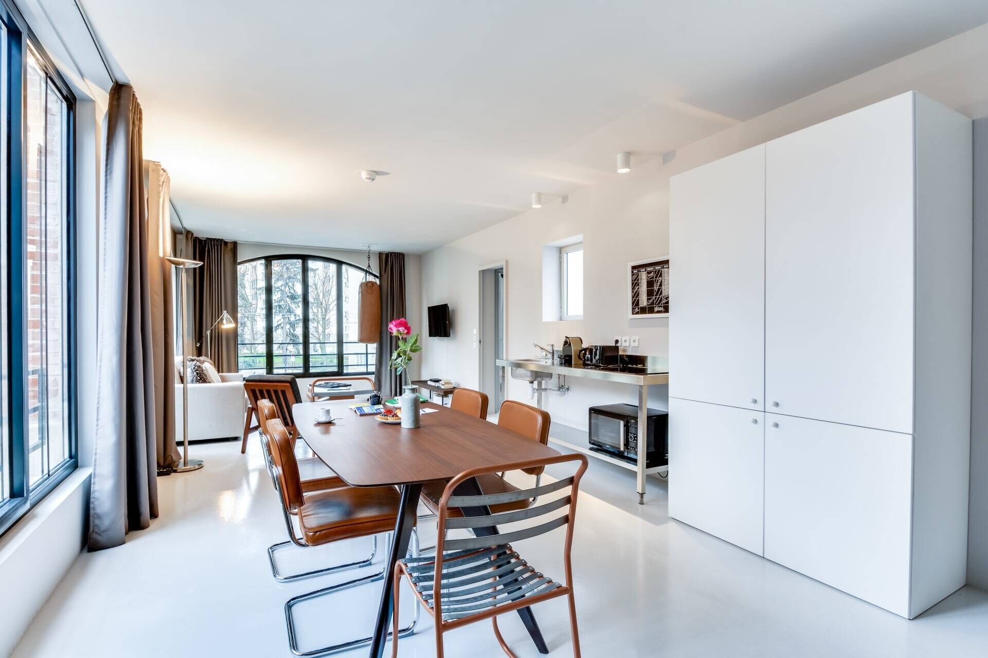 Una cocina moderna con armarios blancos, un comedor con mesa de madera y sillas de metal, y un ventanal que ofrece vistas al exterior.