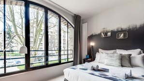 A modern bedroom with a large window, a bed with white and grey bedding, a bedside table with a lamp, and a view of buildings and trees outside. - Sweett | Brune VIII - Three Bedroom Apartment, Sleeps 8 (Paris)