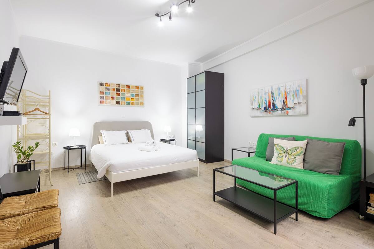 A modern hotel room with a bed, a green sofa, a coffee table, a TV, and a wall art piece.