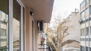 Un balcón con ventanas grandes, piso de baldosas y vista a una calle con edificios y un árbol.