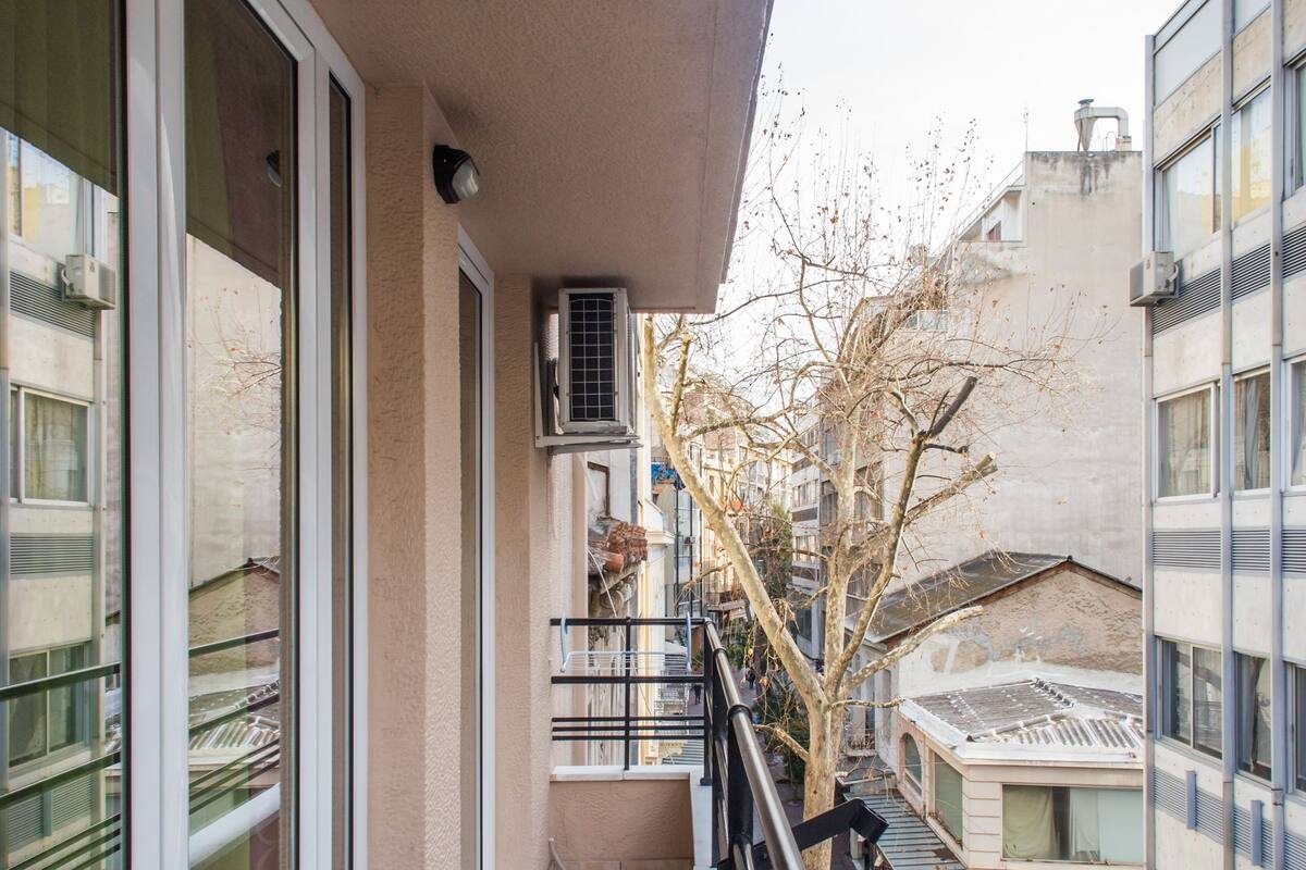 A balcony with large windows, a tiled floor, and a view of a street with buildings and a tree.