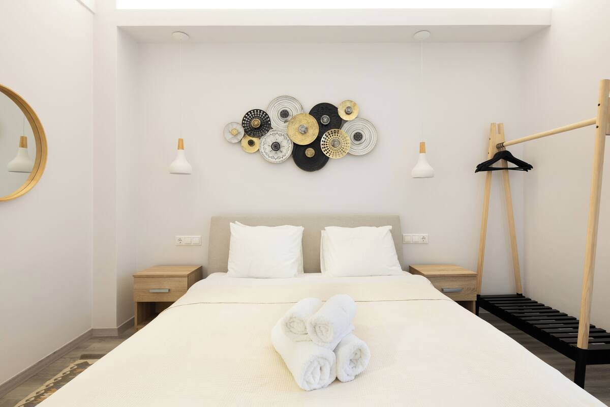 A neatly made bed with rolled towels, a wall-mounted circular light fixture, and a wooden hanger rack.