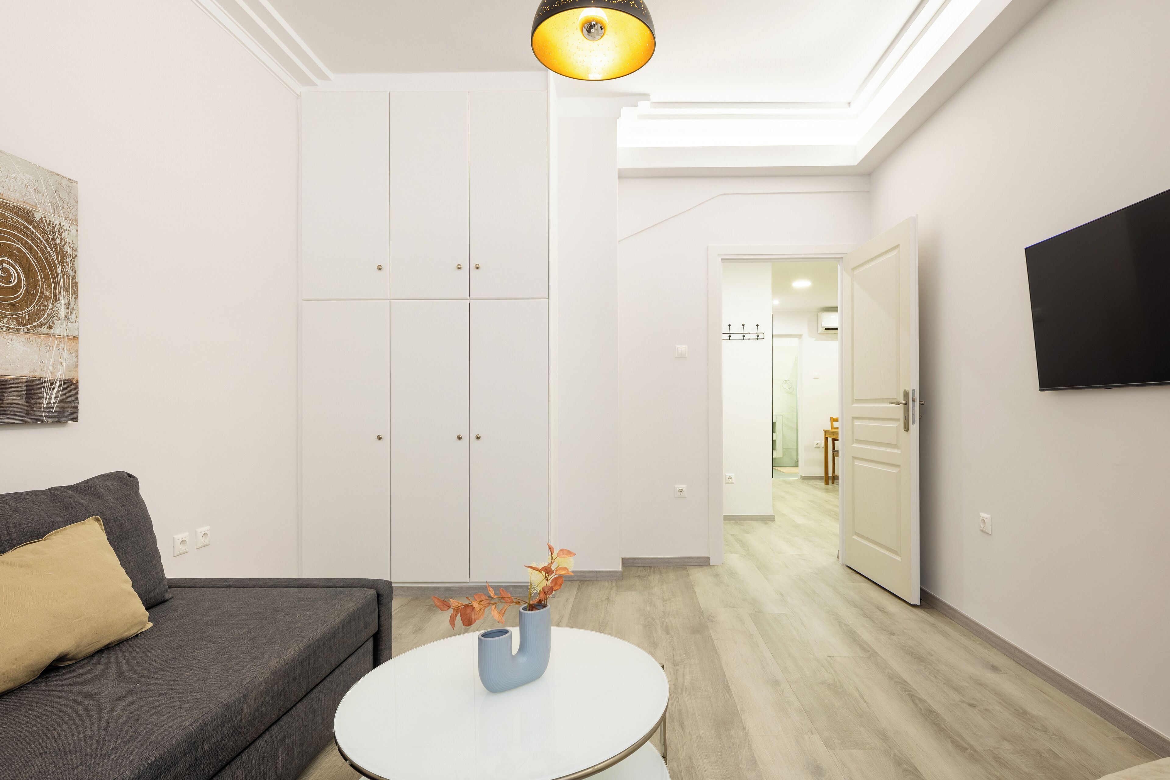 A modern living room with a grey sofa, a white coffee table with a vase, a wardrobe, and a TV mounted on the wall.