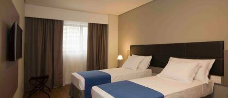 Habitación de hotel con dos camas individuales, un televisor montado en la pared, un escritorio y una ventana con cortinas.