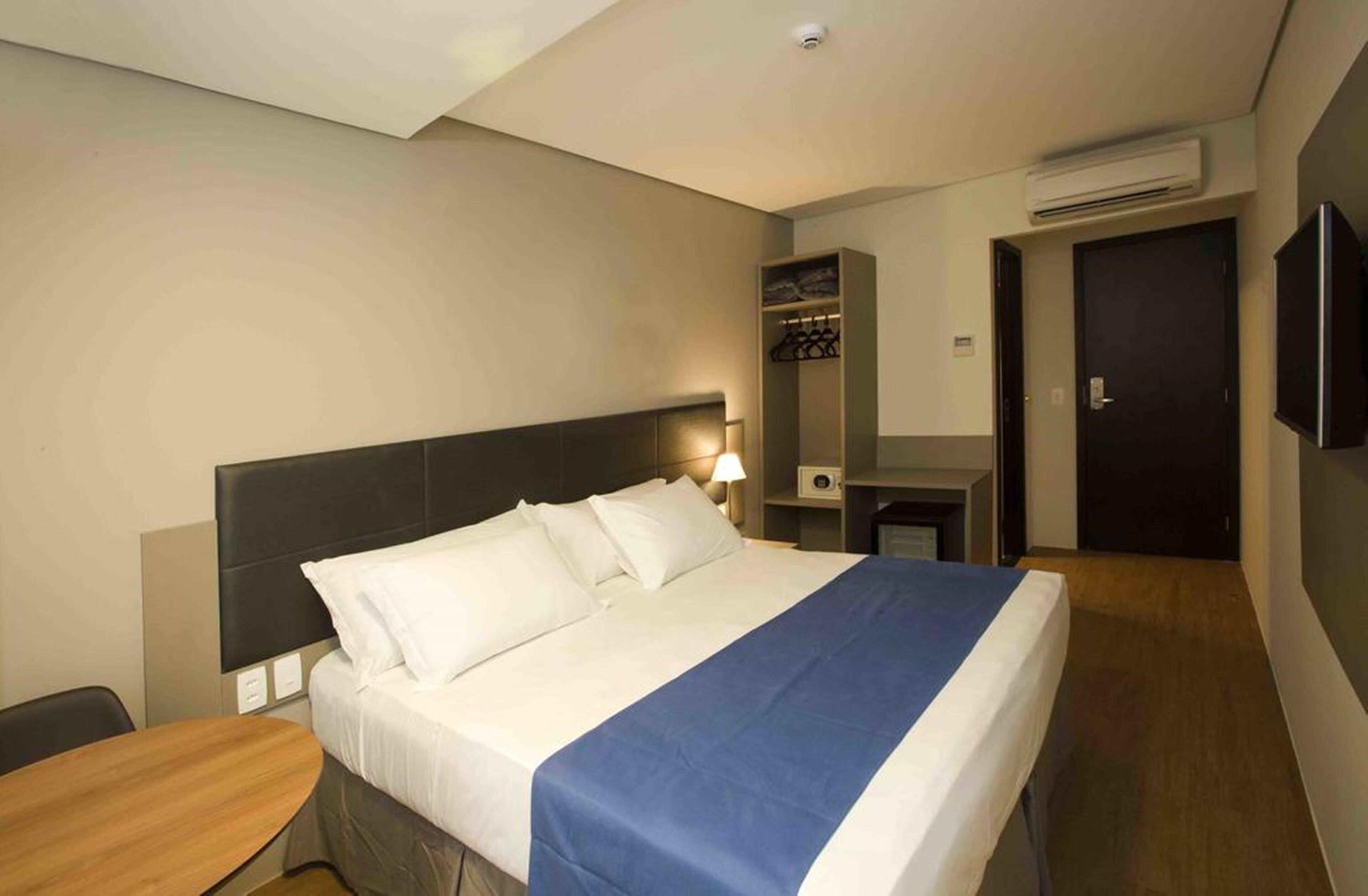 A hotel room with a bed, bedside table, lamp, wardrobe, and a TV mounted on the wall.