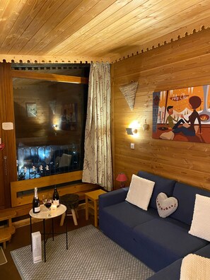 A cozy room with a large window, a blue sofa, a small table with wine bottles, and a painting on the wall.