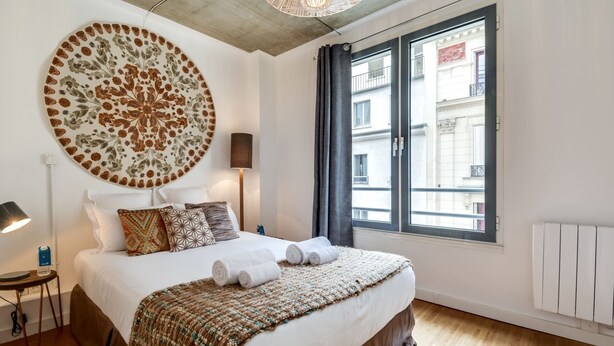 A hotel room with a large bed, a circular floral artwork on the wall, a window with a view of buildings, and a floor lamp. - Sweett | Milan IV - Three Bedroom Apartment, Sleeps 8 (Paris)