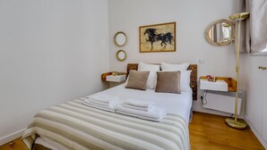 A neatly made bed with white and beige bedding, a painting of a horse on the wall, and a bedside table with a lamp.