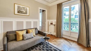 A living room with a grey sofa, a small side table, a radiator, and a window with curtains. - Sweett | Saint Germain IV (Paris)
