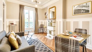 A living room with a sofa, a dining table, a fireplace, and a large window with curtains. - Sweett | Saint Germain IV (Paris)