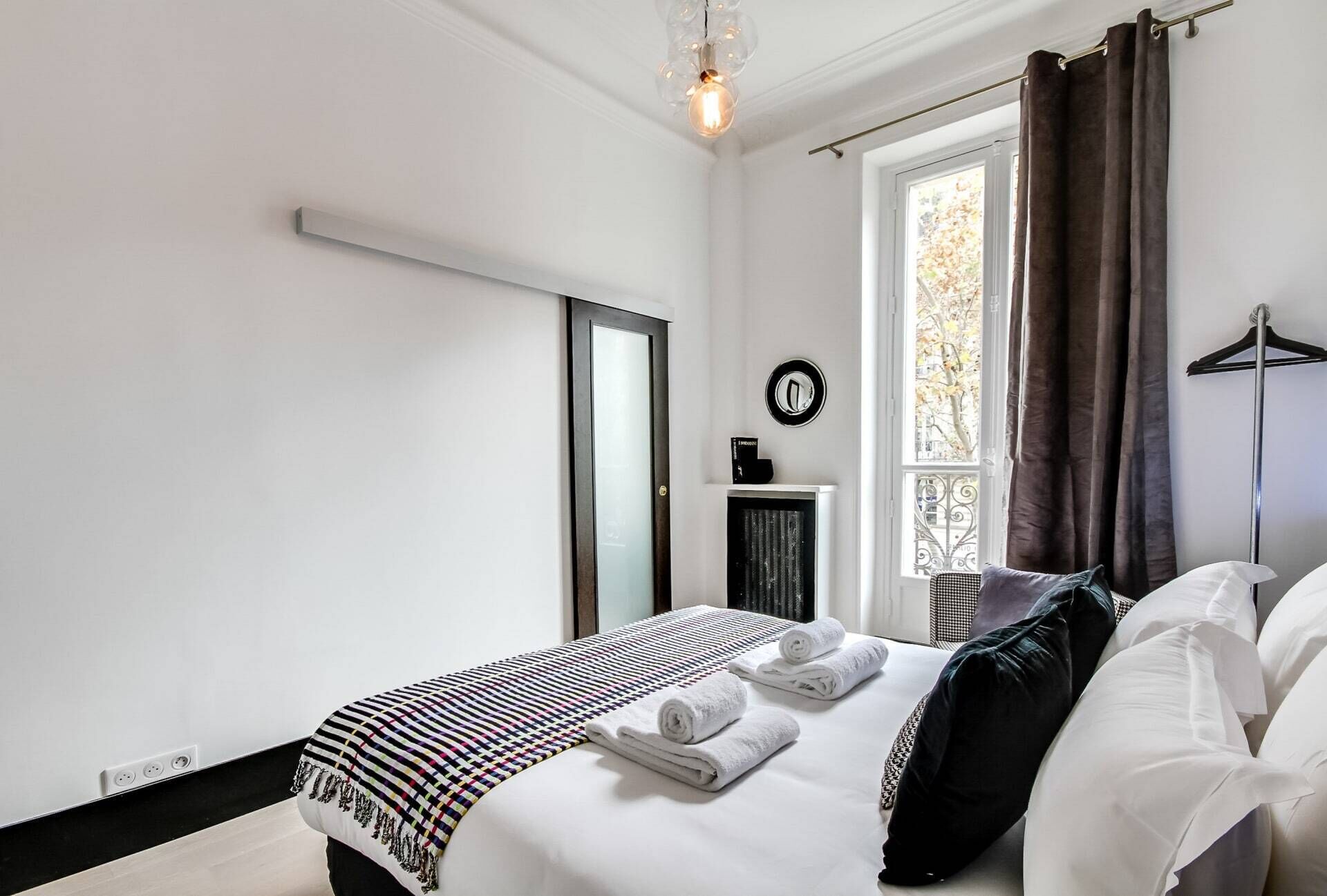 A modern hotel room with a large bed, a window with curtains, a round mirror, and a small side table.