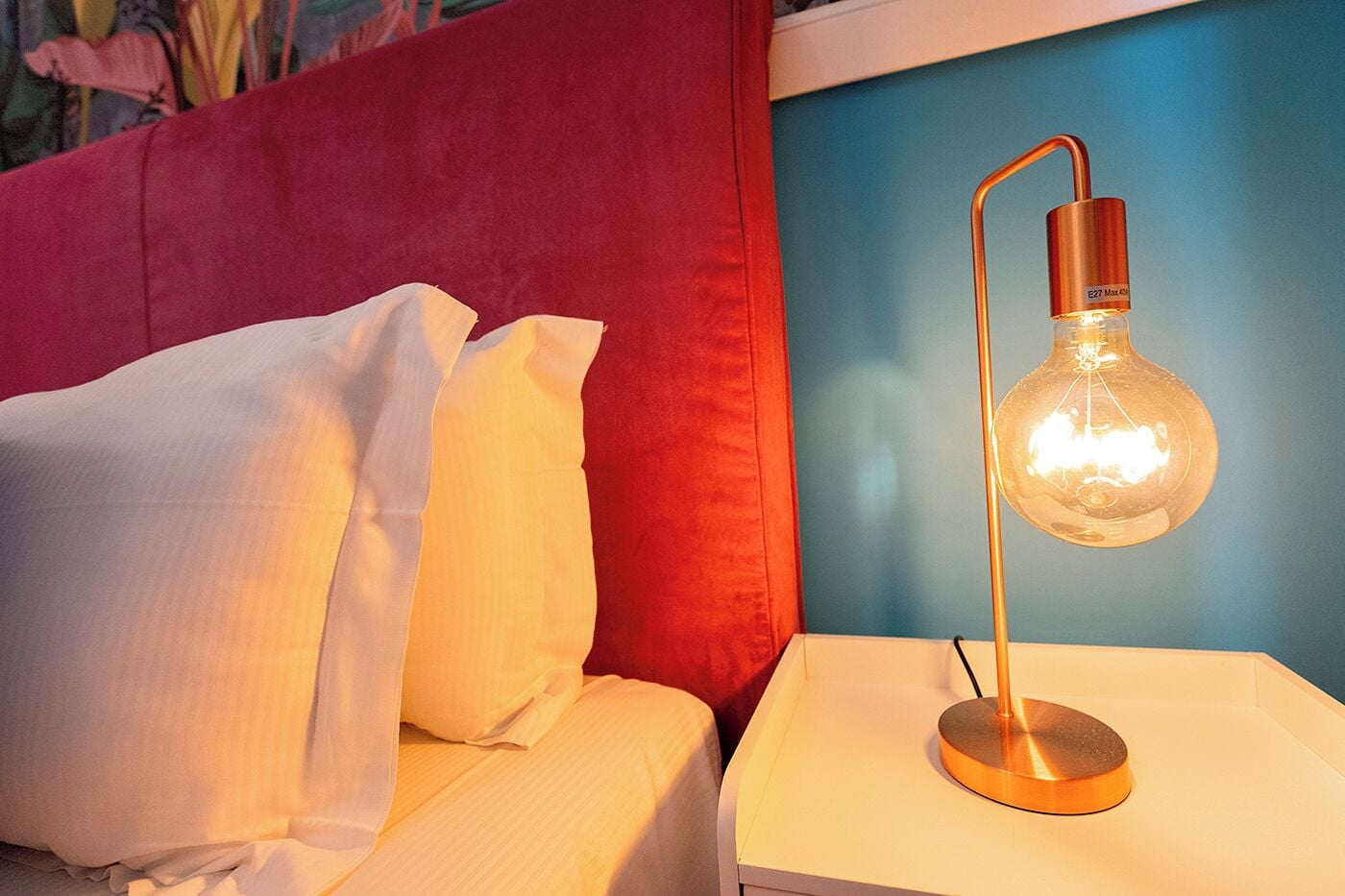 A bed with white bedding and a red headboard, a white nightstand with a modern lamp, and a blue wall in the background.