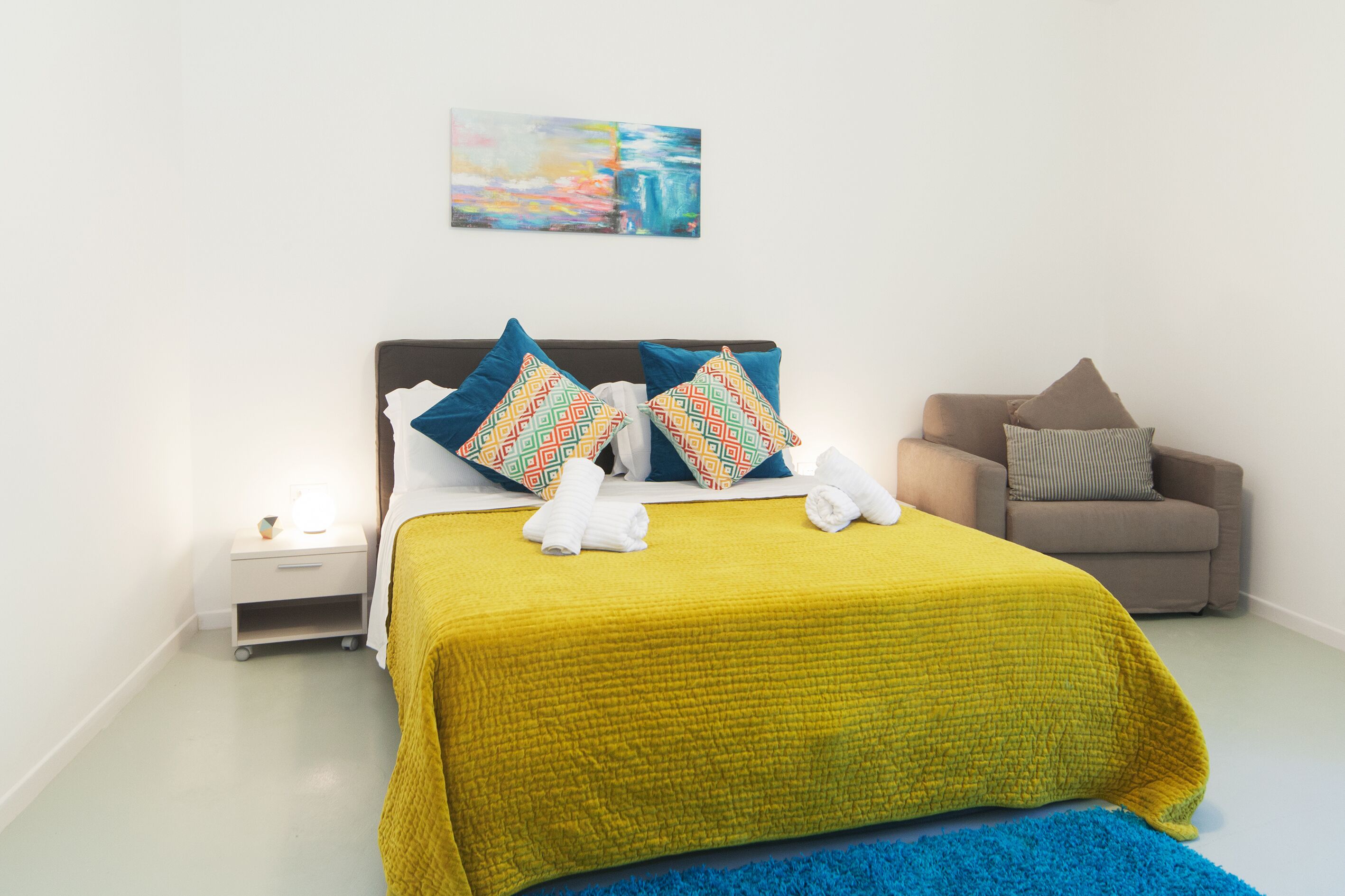 A neatly made bed with a yellow blanket, two patterned pillows, and a blue throw rug.