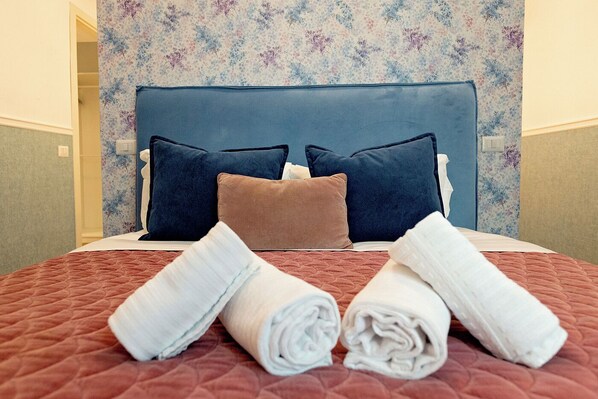 A neatly made bed with a blue headboard, multiple pillows, and rolled towels on a red quilted bedspread.