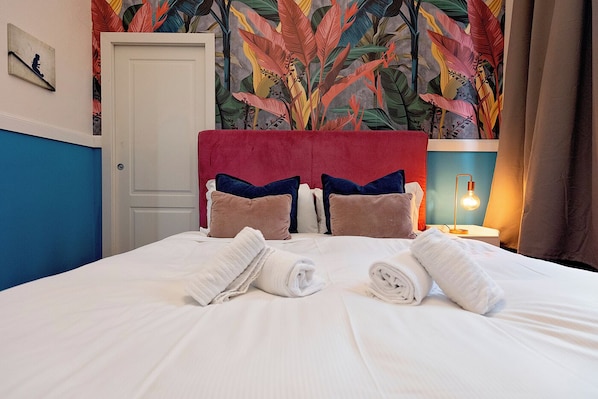 Una cama bien tendida con sábanas blancas y toallas enrolladas, un cabecero rojo y un papel tapiz tropical colorido.