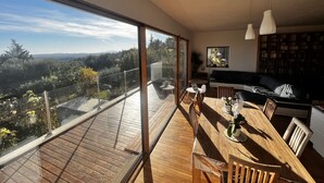 A modern living room with a large sliding glass door leading to a wooden deck, a dining area with a wooden table and chairs, and a cozy seating area with a sofa and bookshelves. - Panoramic Sea View. Take a walk to the medieval village. (Tourrettes-Sur-Loup)