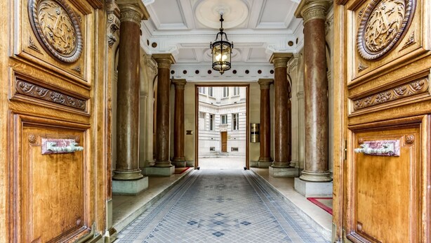 A grand hallway with ornate wooden doors, columns, and a patterned floor leading to a building with multiple windows.