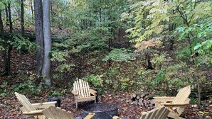 Et skovlandskab med et bålsted og fire træ- Adirondack-stole placeret omkring det.