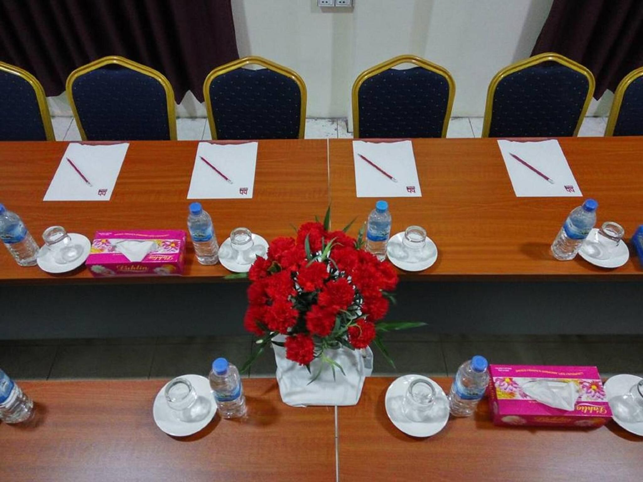 A conference room with a long table, chairs, water bottles, tissues, and a vase of red flowers.