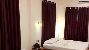 A hotel room with two beds, dark curtains, a small bedside table with a flower arrangement, and a wall-mounted air conditioner.