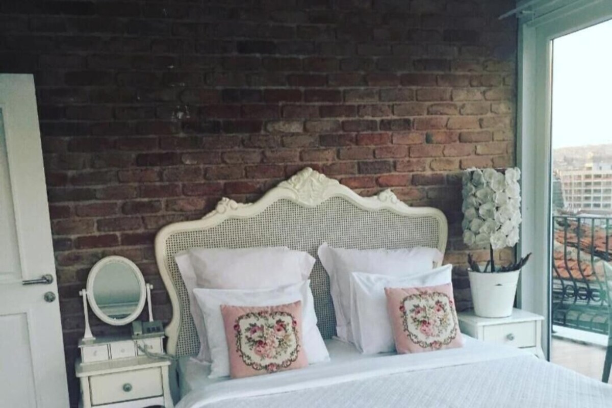 Ein Schlafzimmer mit Backsteinwand, einem weißen Bett mit floralen Kissen, einem Nachttisch mit Spiegel und einer Zimmerpflanze.