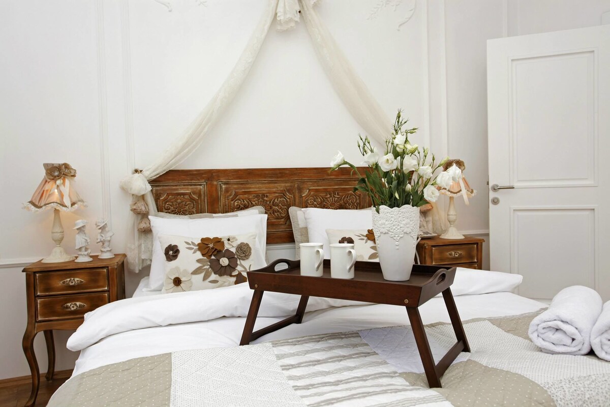 Ein ordentlich bezogenes Bett mit Holz-Kopfstück, ein Nachttisch mit Lampe und ein Tablett mit Blumen.