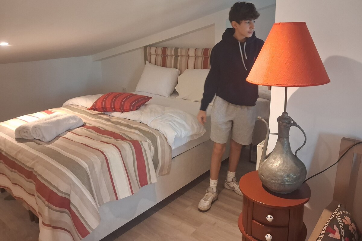 Ein Schlafzimmer mit Bett, Kissen, einer Streifen-Decke, einem aufgerollten Handtuch, einer Lampe und einem kleinen Tisch.