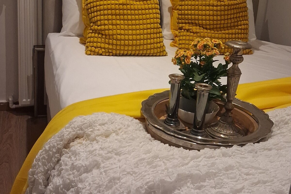 Ein Bett mit weißer Decke und zwei gelben Kissen, ein dekorativer Tablett mit Blumen und eine Wand mit Muster.