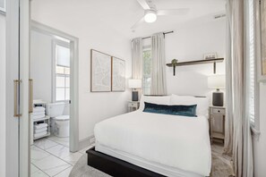 A bedroom with a bed, bedside tables, a ceiling fan, a window with curtains, a wall-mounted shelf with a plant, and a bathroom with a toilet and towel rack.