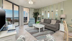 Ein Wohnzimmer mit Sofa, Sesseln, Couchtisch und Fernseher. Ein Balkon bietet Blick auf den Ozean.