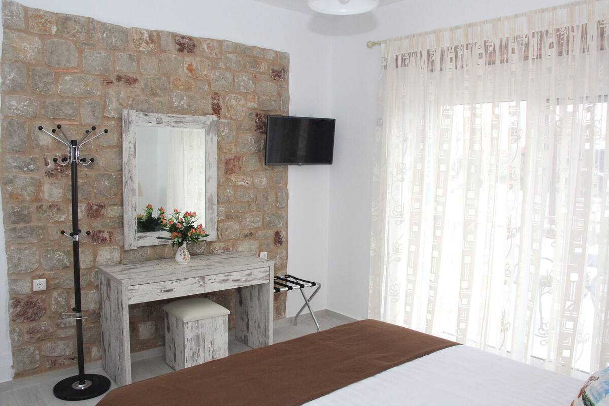 A bedroom with a stone wall, a wooden dresser, a flat-screen TV, and a window with sheer curtains.