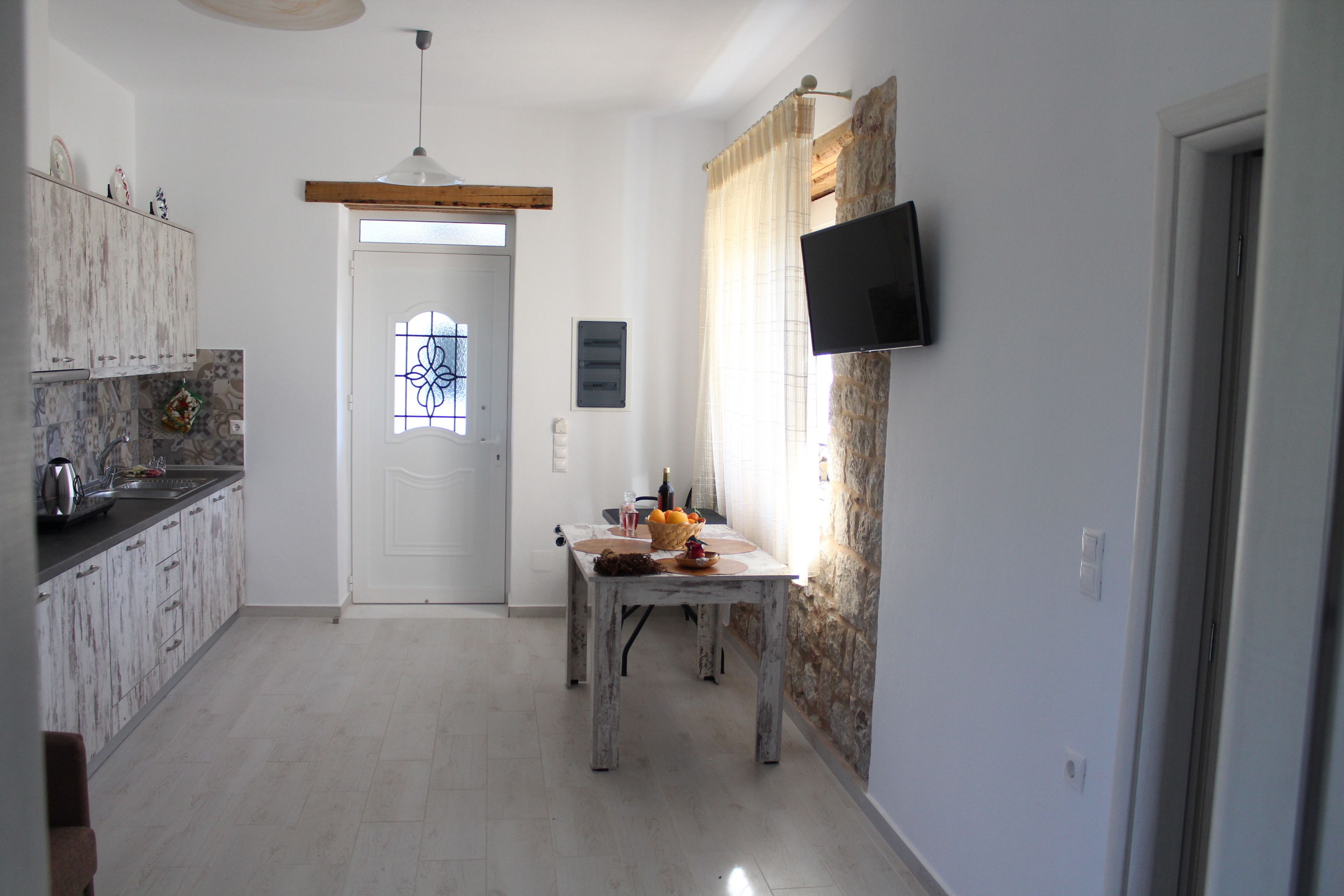 A kitchen with white cabinets, a dining table with a basket of fruit, and a TV mounted on the wall.