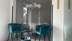 A modern hotel room with a glass door marked "Hotel Ponte," two teal chairs, a small round table, and a teal storage basket.