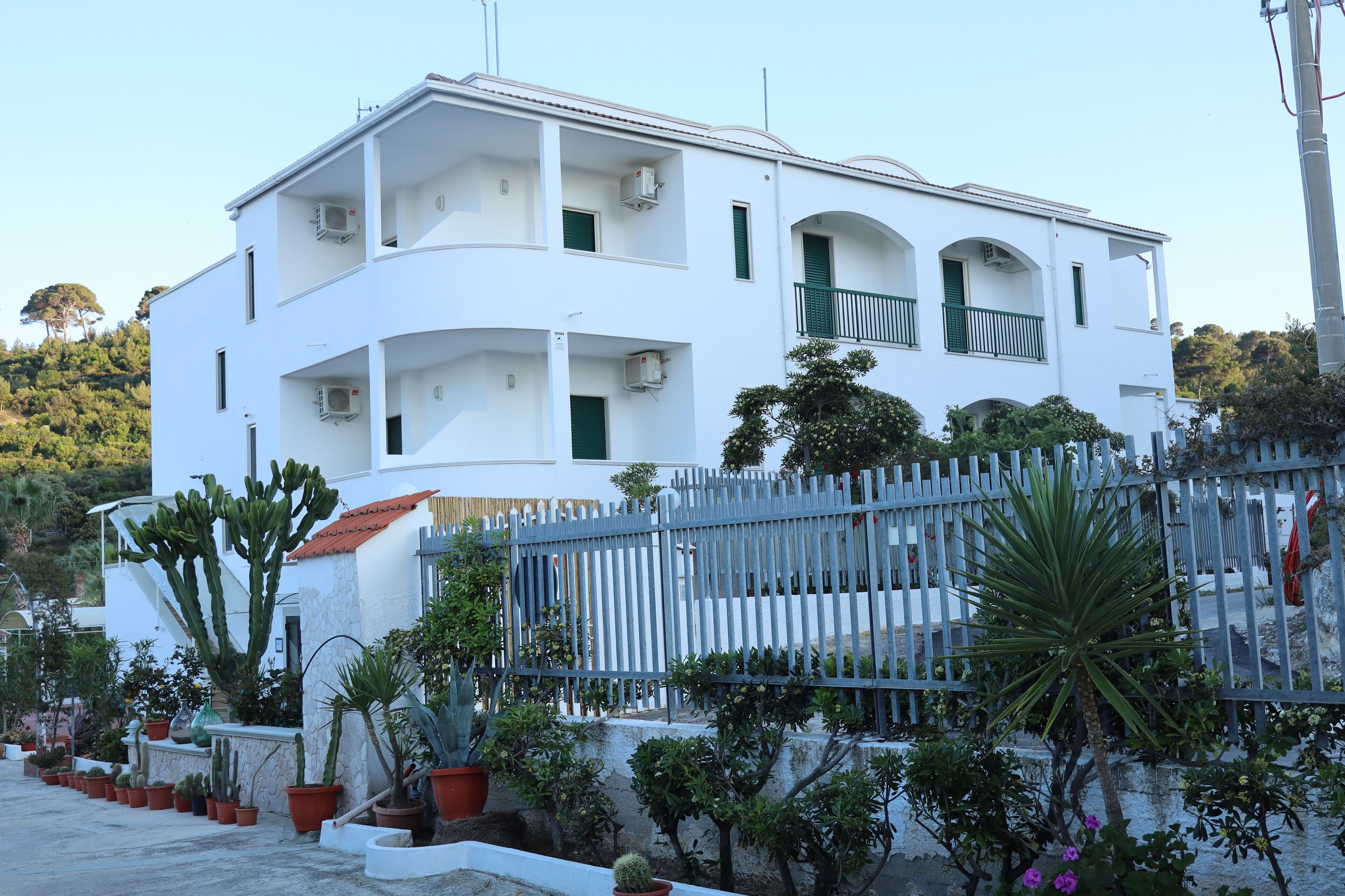 A white multi-story building with balconies, surrounded by potted plants and a fence.
